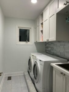 Space-efficient laundry room with white upper cabinets, open shelving, grey tile backsplash, and extended counter over appliances by Polymita Woodcraft
