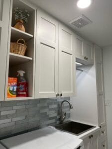 White laundry room cabinets featuring open shelving storage, grey tile backsplash, and undermount sink by Polymita Woodcraft